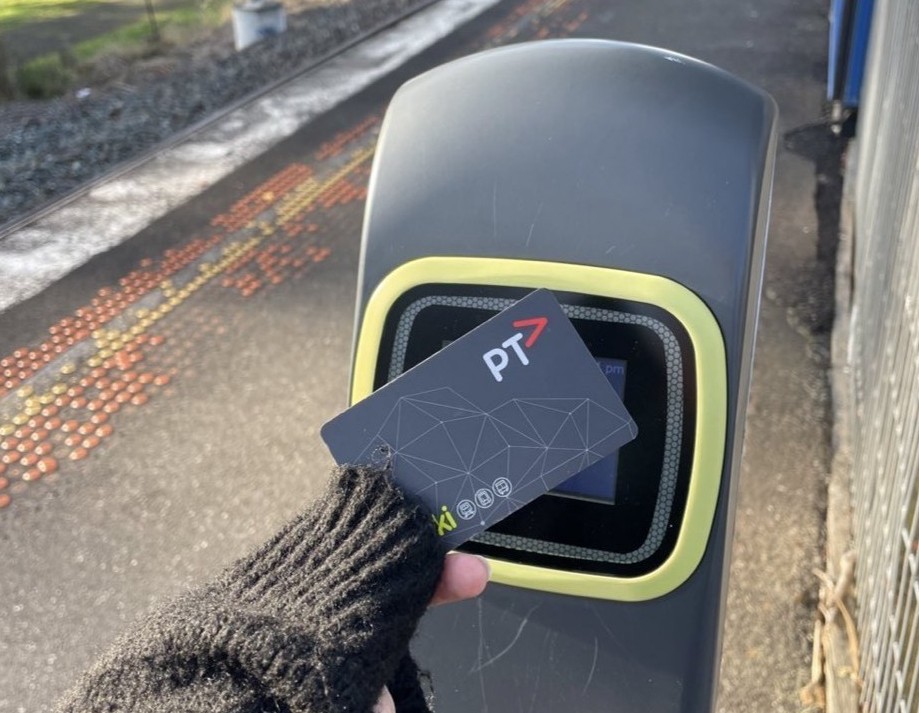 A woman's hand, covered in the sleeve of a black cardigan raches out to touch a myki to a green and grey myki reader. The ashfelt of a train station platform can be seen in the background.